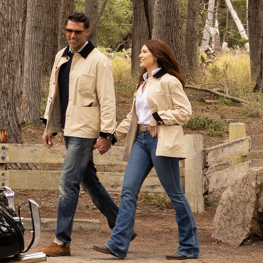 Man and woman walking together in a forested area with a motorcycle nearby.