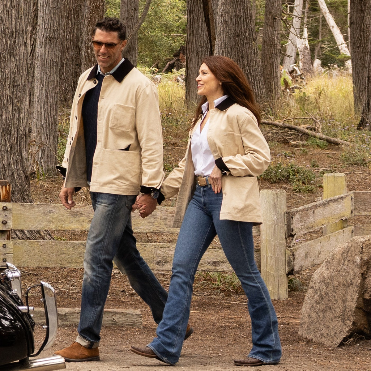 Man and woman walking together in a forested area with a motorcycle nearby.