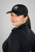 Woman wearing a black cap with a pebble beach logo and jacket against a gray background