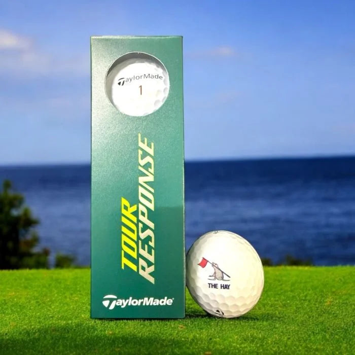 TaylorMade golf ball with The Hay logo and packaging on a grassy area with ocean view